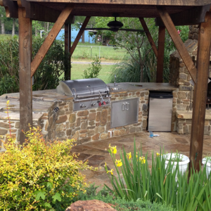 A large backyard patio featuring a stone fireplace and a stone bar with a stainless steel fridge and grill built in.
