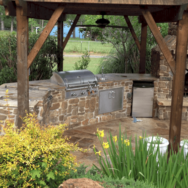 A large backyard patio featuring a stone fireplace and a stone bar with a stainless steel fridge and grill built in.