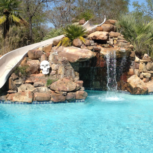 A backyard waterfall next to a slide going down a stone installment leading towards a bright blue pool.