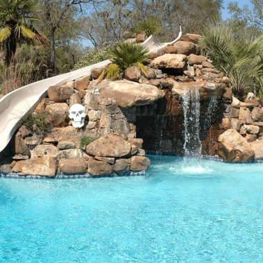 A backyard waterfall next to a slide going down a stone installment leading towards a bright blue pool.