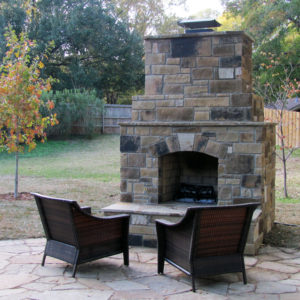 A stone fireplace in a back yard with two chairs facing it.