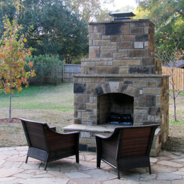 A stone fireplace in a back yard with two chairs facing it.