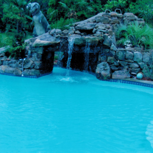 A stone waterfall overhanging a backyard pool, with a statue of a dinosaur in the background.
