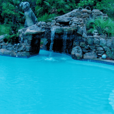 A stone waterfall overhanging a backyard pool, with a statue of a dinosaur in the background.