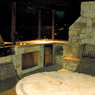A backyard grill in a stone bar with a large stone chimney and steel mini fridge.