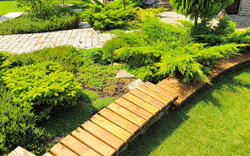 A narrow wooden pathway over a lush green garden, with nicely landscaped trees and a stone pathway in the background.