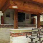 A backyard bar covered with a large wood overhang, with chairs facing a mounted TV.