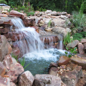 A backyard stone waterfall dumping water into a small pond surrounded by rocks and greenery.
