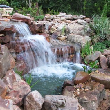 A backyard stone waterfall dumping water into a small pond surrounded by rocks and greenery.