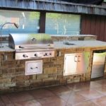 A stainless steel grill and, mini fridge, and sink mounted in a masterfully-crafted stone countertop in a backyard.